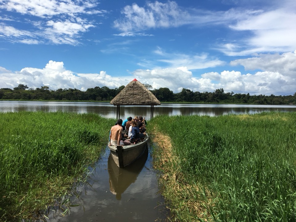 A wooden canoe with people aboard gliding down a reed channel toward a thatched lookout on the lake